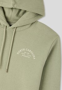 Felpa verde in cotone con cappuccio regolabile. Presenta testo bianco: "NORTH CAROLINA EST 1982 ELITE CLUB WILMINGTON." Texture liscia.