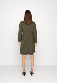 Vestido verde oliva con mangas largas y cintura ajustada, con un dobladillo ligeramente acampanado y tela suave. Se lleva con zapatos de tacón cuadrado negros.