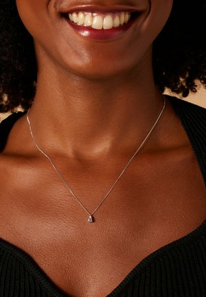 Personne souriante à la peau foncée portant un délicat collier en argent avec un petit pendentif, vêtue d'un haut noir texturé.