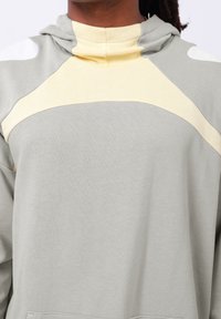 Lys grå hettegenser med myk tekstur, som har et gult og beige geometrisk design på brystet og hette. Ingen synlige detaljer.
