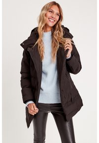 Schwarze Daunenjacke mit Kapuze, mit einem Reißverschluss an der Front und gestepptem Muster. Getragen über einem hellblauen Pullover und schwarzen Hosen.