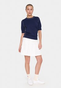 Haut tricot navy à manches courtes avec passepoil blanc, associé à une jupe plissée blanche et des baskets blanches. Des chaussettes neutres complètent le look.