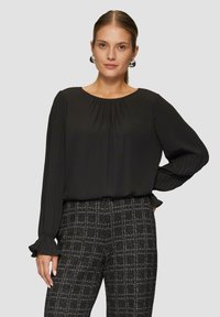 Schwarze Bluse mit langen Ärmeln, gefpleiteten Ärmeln und rundem Halsausschnitt, kombiniert mit strukturierten karierten Hosen in Schwarz und Weiß.