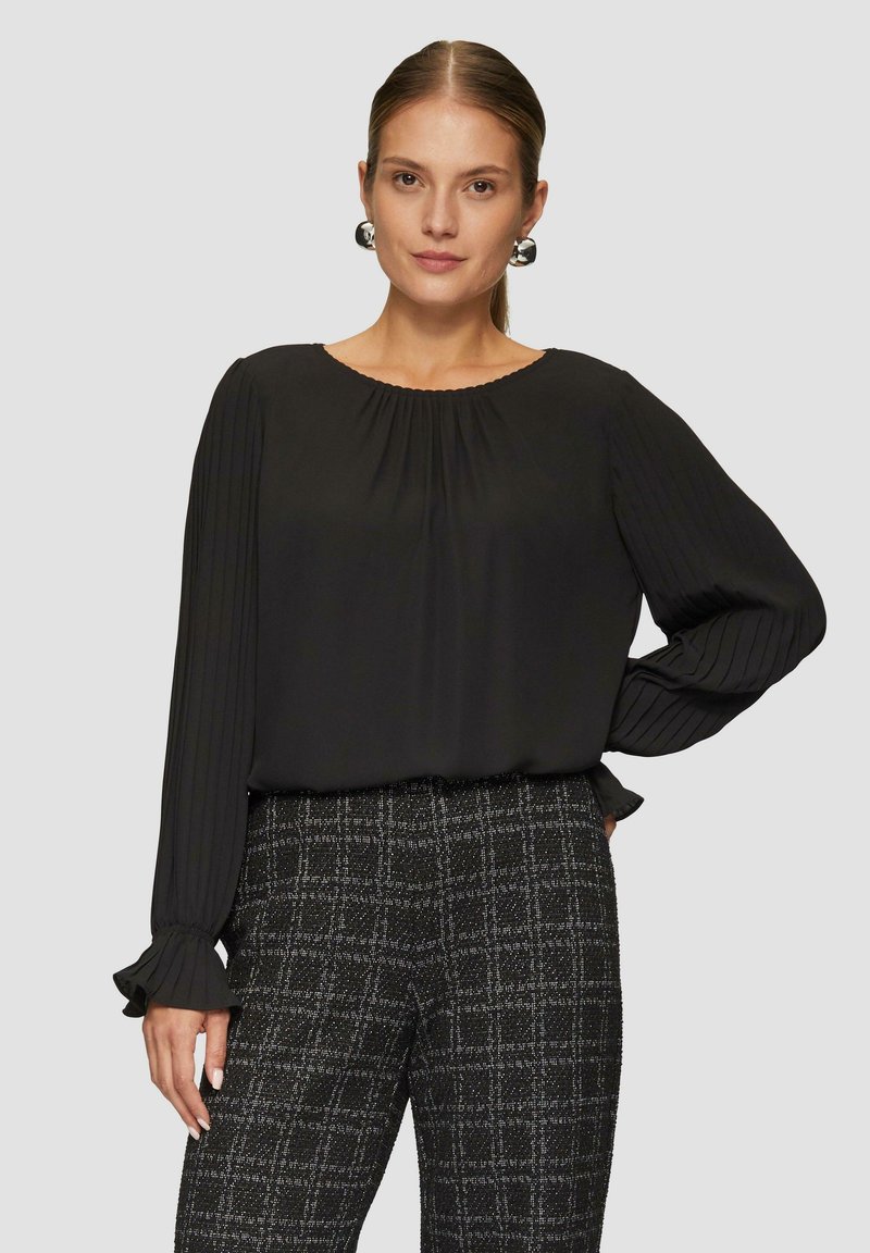 Schwarze Bluse mit langen Ärmeln, gefpleiteten Ärmeln und rundem Halsausschnitt, kombiniert mit strukturierten karierten Hosen in Schwarz und Weiß.