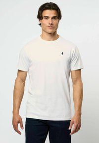 Jauns vīrietis, valkājot vienkāršu baltu īsajām piedurknēm t-kreklu ar nelielu tumšu logotipu kreisajā krūtīs, apģērbts tumšās džinsās, stāv pret vienkāršu fonu.