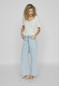 Camiseta de cuello en V gris claro, de corte holgado, combinada con vaqueros de mezclilla azul claro de talle alto y pernera ancha. Modelo lleva puestas unas sandalias blancas de tacón.