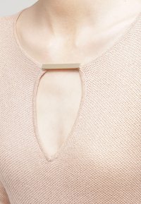 Jasnoróżowy sweter z teksturowanym splotem, z dekoltem w kształcie litery V i złotym prostokątnym akcentem przy dekolcie.