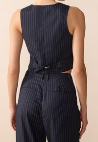 Person klädd i en marinblå ränder utan ärmar väst och matchande hög midja byxor, sedd bakifrån mot en neutral bakgrund.