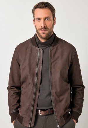 Hombre con cabello corto castaño y barba, vestido con una chaqueta de cuero marrón oscuro sobre un suéter negro de cuello alto, con las manos en los bolsillos, frente a un fondo sencillo.