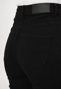 Jeans negros con un diseño ajustado, fabricados en material de denim, con bolsillos traseros y una etiqueta de cuero en la cinturilla. Textura suave.