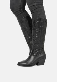 Botas de vaquero de cuero negro con puntas afiladas, caños altos que presentan costuras decorativas en relieve y tacones apilados. Detalle de cremallera en el lateral.
