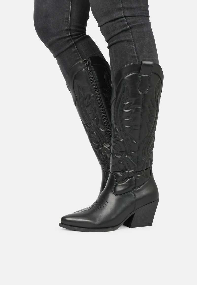 Botas de vaquero de cuero negro con puntas afiladas, caños altos que presentan costuras decorativas en relieve y tacones apilados. Detalle de cremallera en el lateral.