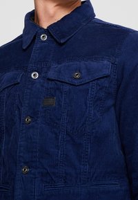 Man bär en mörkblå manchesterjacka med knappade bröstfickor och en liten svart logotypmärke på vänstra sidan.