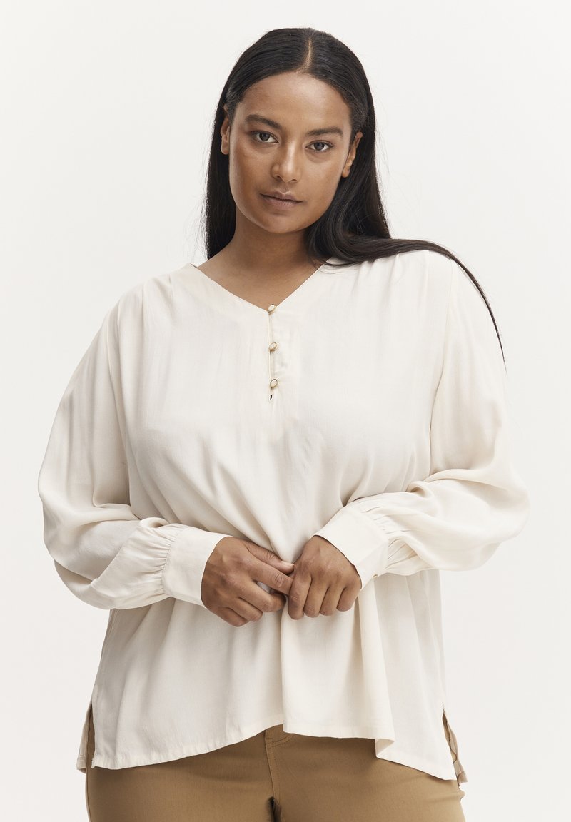 Blusa beige chiaro con scollatura a V, tre bottoni all'collo e maniche lunghe. Presenta spacco laterale e vestibilità ampia. Tessuto liscio.