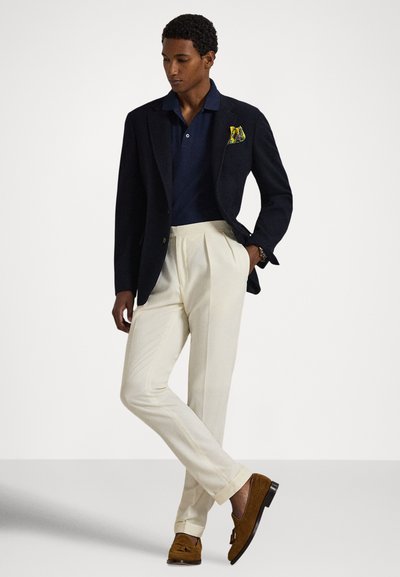 Μπλε ναυτικό πόλο πουκάμισο, σκούρο μπλε σακάκι ραμμένο στα μέτρα, κρεμ πλισέ παντελόνια, καφέ λοφιάδες και ένα τσεμπέρι με σχέδιο, παρουσιασμένα σε ουδέτερο φόντο.