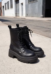 Bottes noires épaisses à plateforme avec lacets et semelles hautes, posées sur un trottoir en béton près d'une rue urbaine.