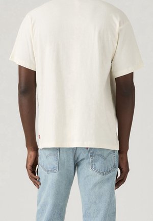Personne portant un t-shirt blanc uni à manches courtes et un jean bleu clair en denim, vue de dos sur un fond uni.