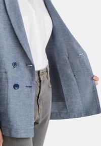 Modrý kostkovaný blazer vyrobený z lehkého materiálu, s dvěma modrými knoflíky a kontrastním šitím. Nosený přes bílé tričko a šedé džíny.