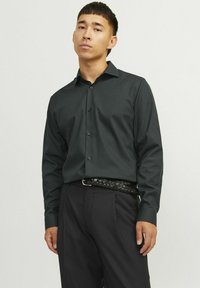Camisa de manga larga verde oscura con cuello abotonado y botones negros. Combinada con pantalones negros y un cinturón trenzado negro.