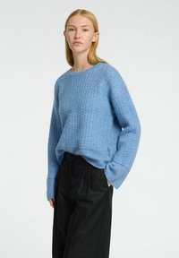 Blå strikket sweater med et tekstureret mønster, rund hals og dropped skuldre, parret med sorte bukser, stående mod en neutral baggrund.