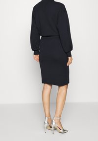 Marinblå stickad klänning med en figurnära silhuett, långa ärmar och ett bälte vid midjan; kombinerad med silverfärgade högklackade skor med ankelsnören.