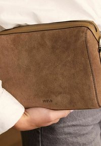 Bruine suede clutch met een soepele leren rits en een subtiele logo aan de zijkant, vastgehouden tegen een wit shirt en grijze spijkerbroek.