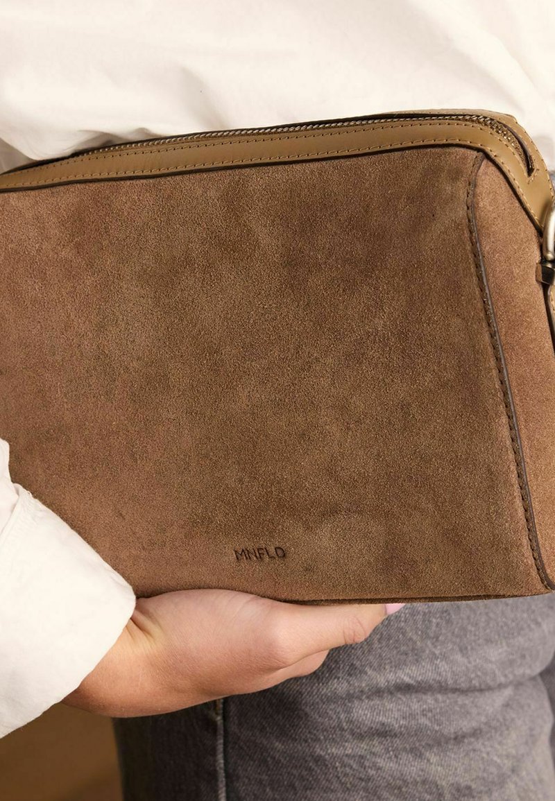 Bruine suede clutch met een soepele leren rits en een subtiele logo aan de zijkant, vastgehouden tegen een wit shirt en grijze spijkerbroek.