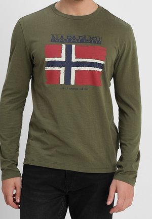 Chemise à manches longues vert olive avec drapeau norvégien usé et logo "NAPAPIJRI" sur la poitrine, portée avec un pantalon noir.