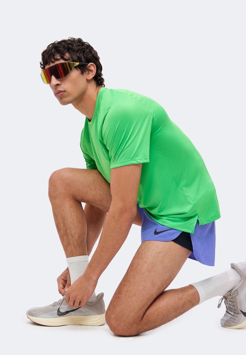 Man wearing a green shirt, blue shorts, and reflective sunglasses kneels to tie his grey running shoe, ready for athletic activity.