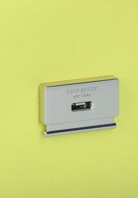 Portacarte metallico in argento, con logo "kate spade" inciso, montato su uno sfondo giallo brillante testurizzato.