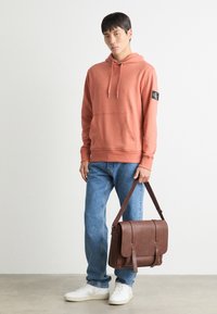Felpa color ruggine con tasca frontale, jeans azzurri chiari, scarpe da ginnastica bianche e una borsa a tracolla in pelle marrone con doppie cinghie.