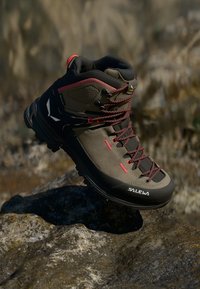 Khaki turistická bota s červenými akcenty, gumovou podrážkou a vysokým kotníkem. Disponuje odolným materiálem a složitým systémem šněrování.