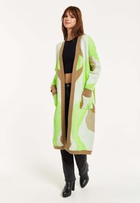 Knit cardigan in green and beige patterns, open front design, long sleeves, two front pockets, with a ribbed beige trim. Black fitted top and pants.