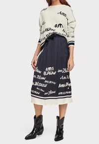 Pull en tricot avec base crème et texte noir. Jupe marine avec écriture blanche et bande inférieure contrastante. Bottines noires.