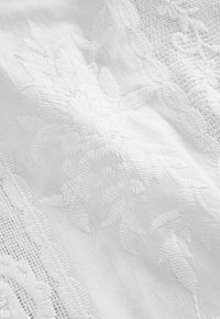 Tissu blanc avec broderies florales détaillées et motifs en dentelle créant des dessins texturés et en relief sur toute la surface.