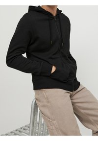 Černá mikina na zip s kapucí, předními kapsami a žebrovanými manžetami. Hladká textilie s kovovým zipem a detaily na šňůrkách.