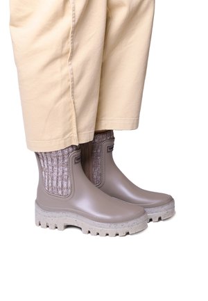 Stivali alla caviglia in gomma marroni con dettaglio in maglia testurizzata, suola spessa e accenti maculati, abbinati a pantaloni larghi beige.
