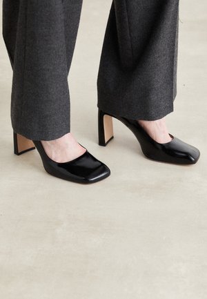 Osoba nosząca czarne wysokie buty na obcasie typu square toe z masywnym obcasem i ciemnoszare spodnie o szerokich nogawkach na jasnej podłodze.