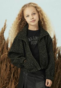 Zielona kurtka z nadrukiem w panterkę na zamek błyskawiczny z elastycznymi mankietami, w komplecie z czarnym t-shirtem z tekstem wysadzanym dżetami oraz czarną skórzaną spódnicą.
