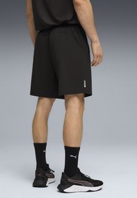 Άνδρας με μαύρο αθλητικό σορτς, μαύρες κάλτσες Puma και μαύρα αθλητικά παπούτσια Puma, στέκεται με την πλάτη προς την κάμερα σε γκρι φόντο.