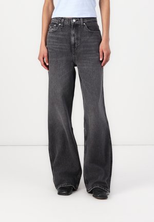 Person iført højtaljede, falmede sorte vidde jeans kombineret med sorte sko og en kort hvid top mod en ensfarvet hvid baggrund.