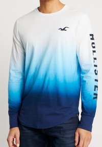 Långärmad T-shirt i vit till blå gradient. Tillverkad av mjukt tyg. Har rund halsringning och tryckt "HOLLISTER" på ärmen.