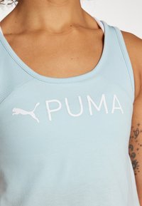 Jasnoniebieski top na ramiączkach wykonany z miękkiego materiału, z białym logo "PUMA" i okrągłym dekoltem. Widać teksturowane panele na ramionach.