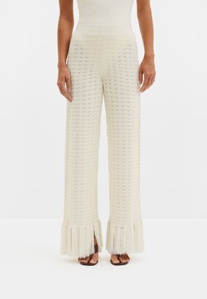 Femme portant un pantalon en maille crème taille haute à jambes larges avec des détails en franges aux ourlets, associé à des sandales noires.