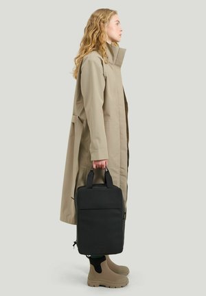 Nainen beigen värisessä trenssitakissa ja saappaissa, pitäen mustaa pystysuoraa suorakulmaista laukkua, seisoo sivuprofiilissa yksiväristä taustaa vasten.