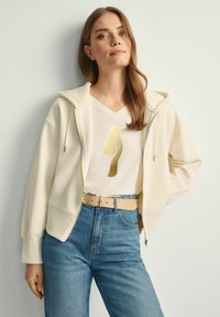 Kremowa bluza z kapturem, zestawiona z białym T-shirtem w serek z złotym wzorem, niebieskimi spodniami z wysokim stanem oraz beżowym paskiem.