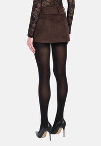 Mini-jupe en daim marron à taille haute avec passants pour ceinture, associée à des collants noirs transparents et des chaussures à talons noires.