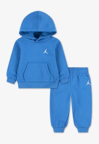 Ensemble sweat à capuche et pantalon bleu en tissu doux. Il présente une poche avant, des poignets côtelés et un logo blanc sur la poitrine et la jambe gauche du pantalon.