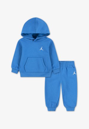 Ensemble sweat à capuche et pantalon bleu en tissu doux. Il présente une poche avant, des poignets côtelés et un logo blanc sur la poitrine et la jambe gauche du pantalon.