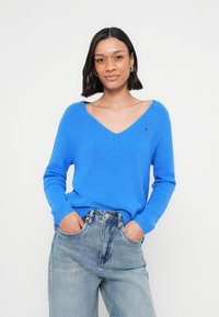 Blue ribbed sweater with a deep V-neck and long sleeves, paired with light blue high-waisted jeans. Subtle logo detail on the chest.